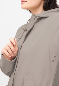 Lichtbeige waterdichte jas met een capuchon, verstelbare trekkoorden, twee zijzakken en drukknoopaccenten. Gladde, matte afwerking.