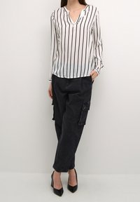 Blusa bianca con strisce verticali nere, scollatura a V, maniche lunghe; abbinata a pantaloni cargo neri larghi con tasche laterali.