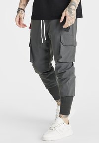 Pantalones cargo de tela gris oscuro con puños ajustados, que incluyen bolsillos laterales, cinturilla elástica y cordones blancos. Combinados con zapatillas blancas.