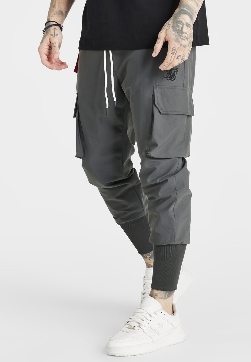 Pantalones cargo de tela gris oscuro con puños ajustados, que incluyen bolsillos laterales, cinturilla elástica y cordones blancos. Combinados con zapatillas blancas.