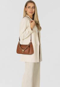 Bolso de cuero marrón con detalle de cadena dorada, forma estructurada y bolsillo frontal, llevado en el hombro sobre un fondo claro.