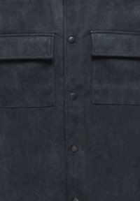 Tumšs zamšādas žakets ar divām krūšu kabatām, pogu aizdari, gludu tekstūru un matētu virsmu. Akcentēts ar melnas krāsas aparatūru.