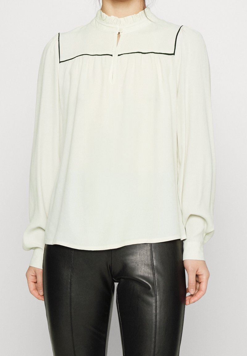 Femme portant un chemisier blanc à manches longues avec un passepoil noir sur le plastron et un pantalon en cuir noir.