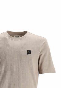 T-shirt beige clair à manches courtes avec des rayures diagonales texturées, un col rond, et un patch carré noir avec un logo sur le côté gauche de la poitrine.