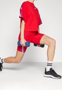 Une personne en tenue de sport rouge effectue des fentes tout en tenant des haltères bleus, portant des baskets noires et des chaussettes blanches avec des rayures noires.