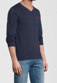 Man in een marineblauwe V-hals trui met een rode geborduurde logo op de borst en lichte spijkerbroek tegen een effen achtergrond.