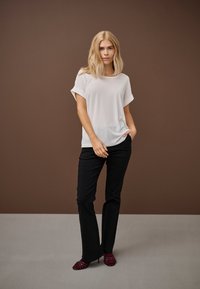 Blonde sieviete valkā baltu brīvu īsroku krekliņu, melnas bikses un burgundijas krāsas augstpapēžu kurpes, stāvot ar vienu roku kabatā pret brūnu fonu.