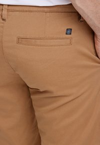 Vaaleanruskeat chinot ovat valmistettu sileästä, tekstuuripinnoitteisesta kankaasta, niissä on tavallinen takatasku ja pieni musta logotagi vyötärön läheisyydessä.