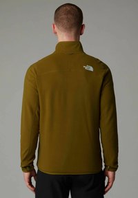 Man seen from back wearing olive green North Face fleece jacket with logo on right shoulder, standing against plain light gray background.