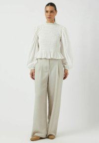 Haut peplum blanc à manches longues avec des détails de smocks et un col haut, associé à un pantalon large plissé gris clair et des chaussures beiges.