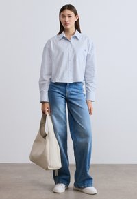 Ριγέ πουκάμισο με κουμπιά σε ανοιχτό μπλε χρώμα, cropped σχέδιο, συνδυασμένο με τζιν ψηλόμεσα μπλε. Καθημερινά λευκά αθλητικά παπούτσια και μια μεγάλη μπεζ τσάντα ώμου.