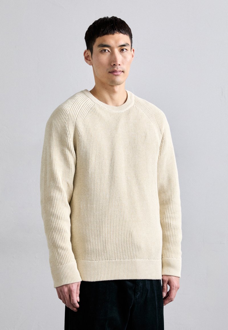 Beige ribbed knit sweater with rounded neckline and long sleeves, featuring a textured fabric and a relaxed fit.