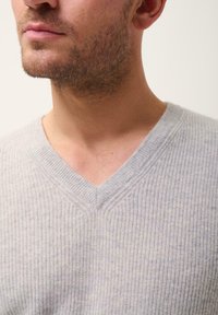 Suéter gris claro de cuello en V con textura acanalada, que presenta un diseño ajustado y sutiles detalles de costura en el escote. Fondo liso.