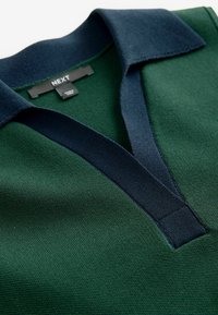 Polo vert avec col marine, texture tricotée et patte de boutonnage. Étiquette visible indiquant "NEXT." Zoom sur les détails du col et du tissu.