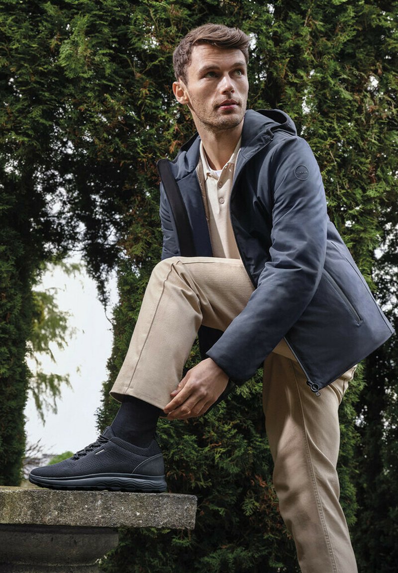 Giacca blu navy resistente all'acqua con cappuccio, pantaloni beige e sneakers nere. La giacca ha una chiusura con zip ed è realizzata in materiale liscio.