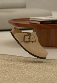 Μπεζ παπούτσι τύπου slip-on με μαύρη αγκράφα, πάτο από φελλό και μαύρη σόλα, αιωρούμενο πάνω από υφασμένο χαλί σε σαλόνι.