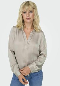 Blouse en soie couleur taupe clair avec un col en V, des manches longues et des poignets boutonnés. Texture lisse et léger éclat. Assortie avec un jean bleu.