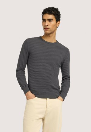 Mężczyzna o kręconych włosach, ubrany w dopasowaną, ciemnoszarą teksturowaną koszulę z długim rękawem i beżowe spodnie, z rękami w kieszeniach, patrzący na bok.