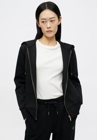 Μαύρο φούτερ με φερμουάρ και κορδόνια, κατασκευασμένο από μαλακό ύφασμα; φορεμένο πάνω από ένα άσπρο ριμπ μπλουζάκι. Διαθέτει χρυσές λεπτομέρειες και χαλαρή εφαρμογή.