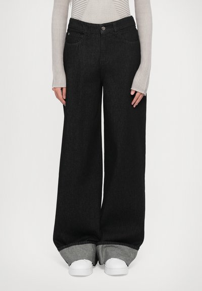 Jeans en denim noirs à jambes larges avec une taille haute, dotés de deux poches avant et de poignets gris repliés, associés à des baskets blanches.