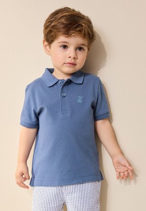 Băiat tânăr cu păr brun deschis, purtând un polo albastru și pantaloni scurți drepți deschiși cu dungi, stând în fața unui perete bej.