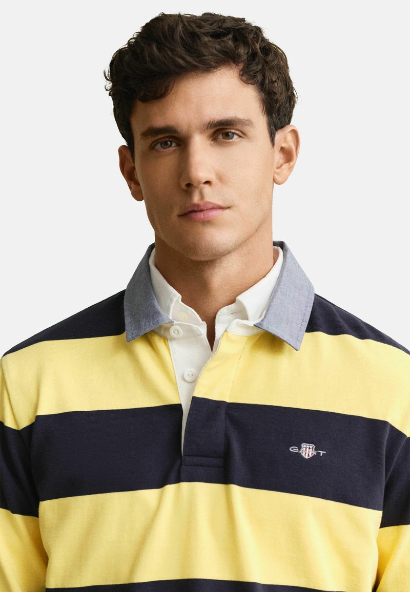 Junger Mann mit lockigem dunklem Haar, der ein gelb- und marineblau gestreiftes Rugbyhemd mit grauem Kragen und weißem Hemd darunter trägt.