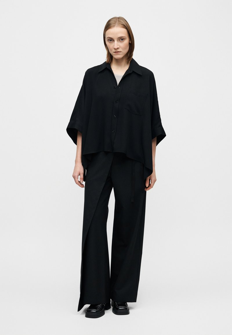 Chemise noire surdimensionnée à manches courtes, fermeture à boutons et poche poitrine, associée à un pantalon noir à jambes larges avec une fente latérale.