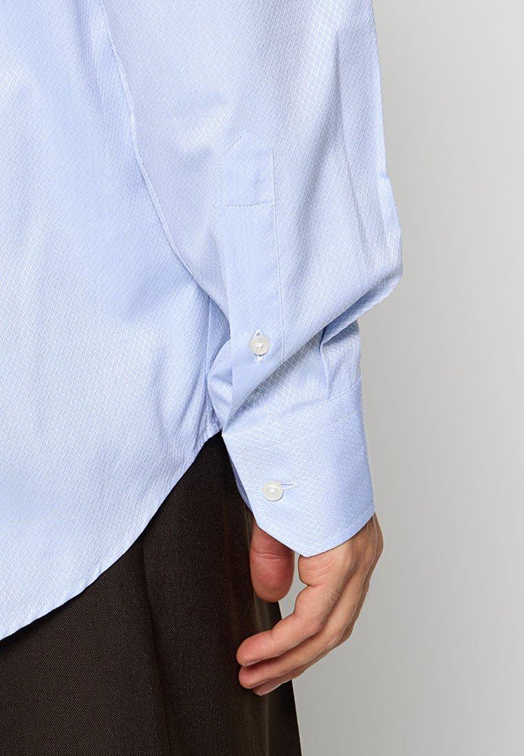 Lichtblauwe shirt met patroon en een gestructureerde afwerking, met een knoopmanchet en een patchzak op de mouw. Donkere broek zichtbaar.