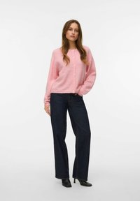 Ροζ ανοιχτόχρωμο πλεκτό ζακέτα με κουμπιά μπροστά, χαλαρή εφαρμογή και ριμπ τελείωμα, φορεμένη πάνω από σκούρο τζιν με φαρδιά πόδια και μαύρα μυτερά μποτάκια.