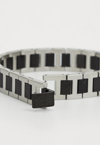 Metallarmbånd med vekslende sølvlenker og teksturerte svarte detaljer, med en svart lås som viser et krokodillelogo.