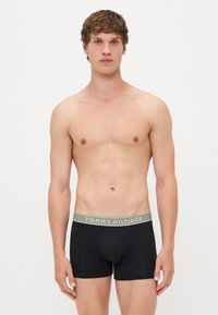 Jovem com cabelo curto e encaracolado está de frente, vestindo cuecas boxer pretas com cintura cinza rotulada "Tommy Hilfiger".