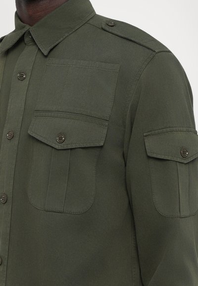 Olivgrünes Button-Up-Hemd mit zwei Brusttaschen und Schulterklappen. Hergestellt aus einem strukturierten Stoff mit dunklen Knöpfen.