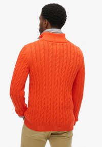 Pomarańczowy sweter z dzianiny warkoczowej z wysokim kołnierzem oraz prążkowanym dołem i mankietami. Pokazany od tyłu, ukazujący teksturowany wzór i dopasowany krój.