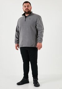 Grå fleece pullover med høj krave, halv lynlås og logo på brystet, parret med sorte tætsiddende bukser og sorte sko.