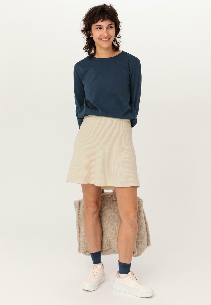 Marineblaues Langarmshirt, beiger gerippter ausgestellter Rock, weiße Sneaker und blaue Socken. Hält eine flauschige beigefarbene Tragetasche. Einfaches und lässiges Design.
