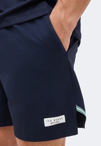 Κοντινό πλάνο ατόμου που φοράει σορτς σε ναυτικό μπλε χρώμα με το χέρι στην τσέπη και το λογότυπο Ted Baker Sport στο τελείωμα.