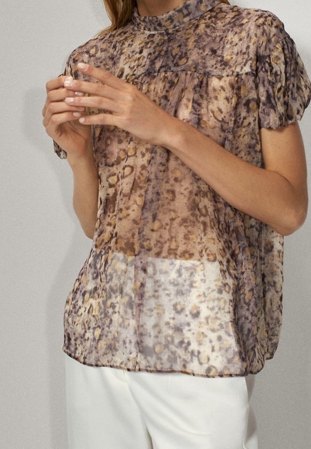 Femme portant un chemisier transparent à manches courtes imprimé léopard avec un col montant et un pantalon blanc, mains jointes doucement devant elle.