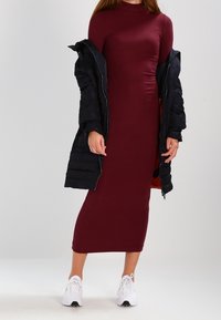 Vestido midi burdeos de canalé con cuello alto, combinado con un abrigo acolchado negro y zapatillas deportivas blancas. Textura suave y diseño ajustado.