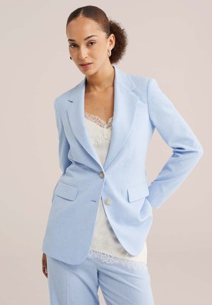 Žena s kučeravými vlasmi v svetlomodrom saku a nohaviciach, na sebe biely top s čipkovým lemom, stojaca na jednofarebnom pozadí.