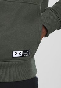 Sudadera verde oliva con puños acanalados, que presenta un parche del logo "Under Armour" en blanco y negro en la parte inferior izquierda. Material suave, corte informal.