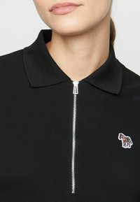 Gros plan d’une personne portant un polo noir avec une fermeture éclair argentée et un petit logo zèbre rayé coloré sur la poitrine.