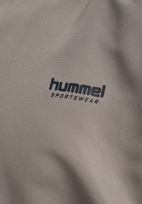 Gråt sportstøjsstof med en tekstureret overflade og en subtil glans. En sort logo med teksten "hummel SPORTSWEAR" er tydeligt synlig.