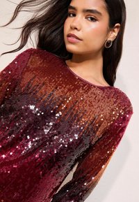 Spīdīga blūze ar sekvīnu, kas pāriet no sarkanās uz purpursarkano krāsu, ar garām piedurknēm un apaļu kakla izgriezumu, uzsverot dzīvīgas un spīdīgas tekstūras.