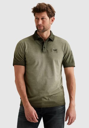 Homme aux cheveux bruns courts et légère barbe, portant un polo vert olive et un jean foncé, regardant vers sa gauche.