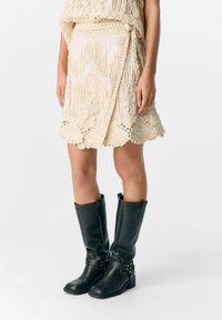 Beige Wickelrock mit aufwendiger, strukturierter Stickerei und gewelltem Saum, kombiniert mit knielangen schwarzen Lederstiefeln mit Schnallen.