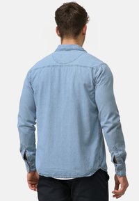 Hombre con una camisa de mezclilla de manga larga azul claro y pantalones negros, visto desde atrás sobre un fondo blanco liso.