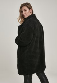 Čierny oversized kabát vyrobený z plyšového materiálu, s širokým golierom, veľkými vreckami a mierne dlhším zadným lemom pre voľný strih.