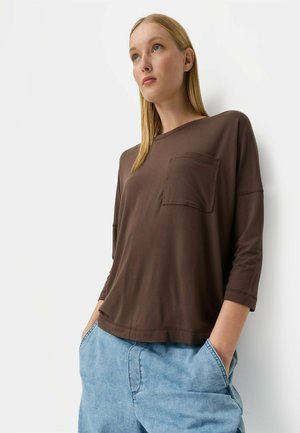 Woman with straight blonde hair wearing a loose brown long-sleeve shirt with a chest pocket and light blue denim pants with hands in pockets.