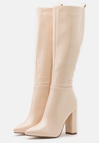 Botines de tacón alto beige hechos de material liofilizado brillante, con punta afilada y tacón cuadrado grueso. Textura suave con cierre de cremallera.
