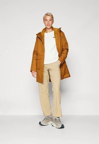 Pessoa em pé, vestindo um casaco com capuz castanho, camisa branca, calças bege e sapatilhas cinzas, contra um fundo claro e liso.